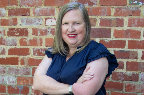 Professional headshot of Emma Olivier standing with arms crossed against a rustic brick wall, wearing a navy blue top and smiling confidently.RetryClaude can make mistakes. Please double-check responses.